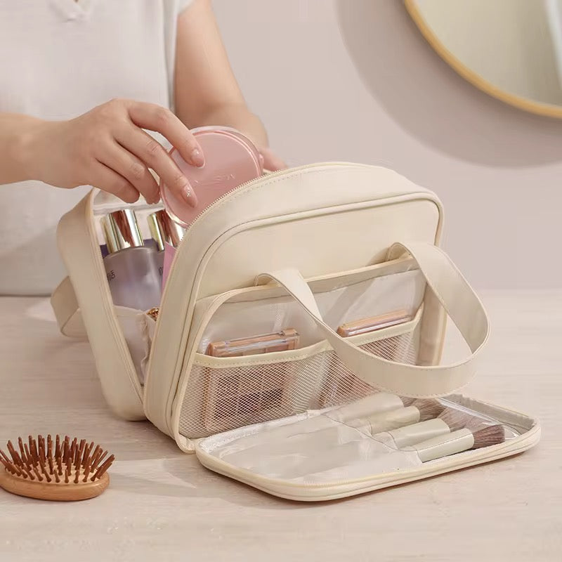 Beige travel makeup bag with compartments, pink compact mirror, and hairbrush on a light surface.