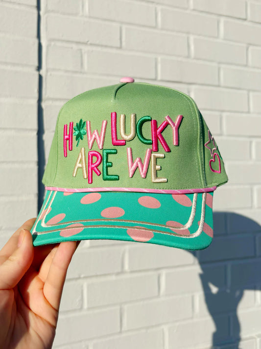 Green cap with colorful text 'HOW LUCKY ARE WE' held against a white brick wall.