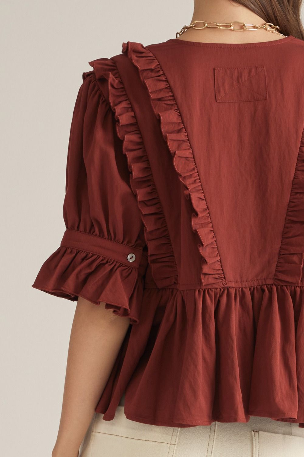 A close-up back view of a woman wearing a rust-brown short-sleeve blouse with ruffled trim and a peplum hem, paired with light beige pants and a gold chain necklace.