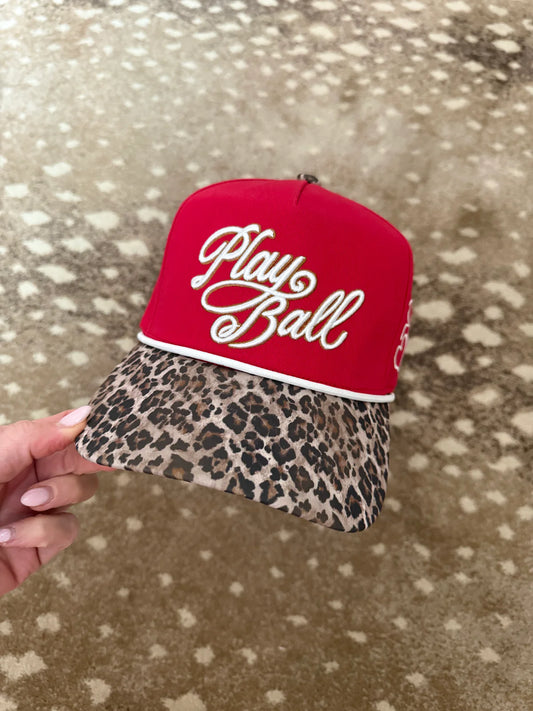 Red cap with 'Play Ball' text and leopard print brim held against a patterned background
