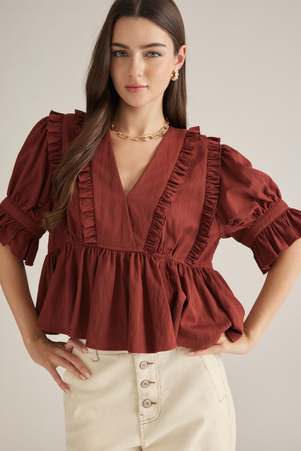 A woman wearing a rust-brown ruffled blouse with puff sleeves and a gathered peplum hem, paired with cream high-waisted pants, against a light beige studio background.