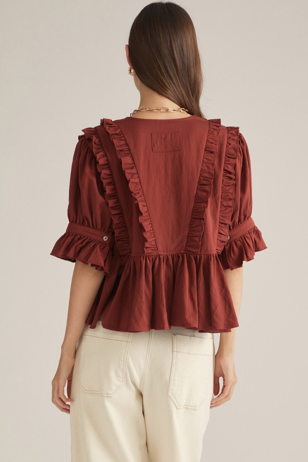 A woman shown from the back wearing a rust-brown ruffled blouse with puff sleeves and cream-colored pants, set against a plain light beige studio background.