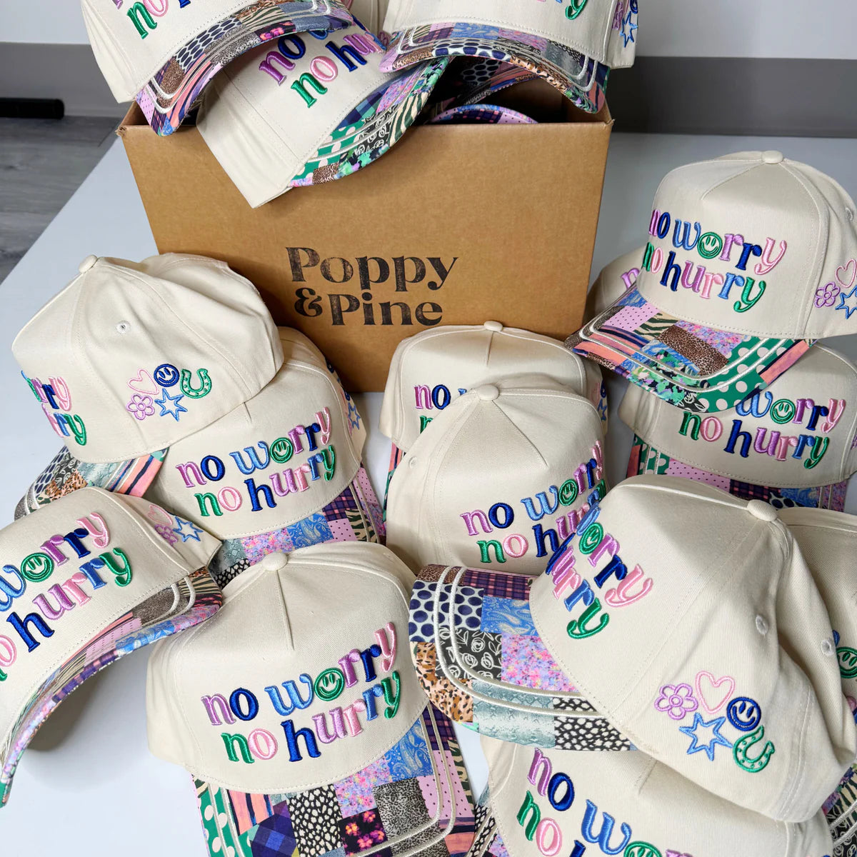 White hats with 'no worry no hurry' text and colorful designs on a white surface with a Poppy & Pine box.