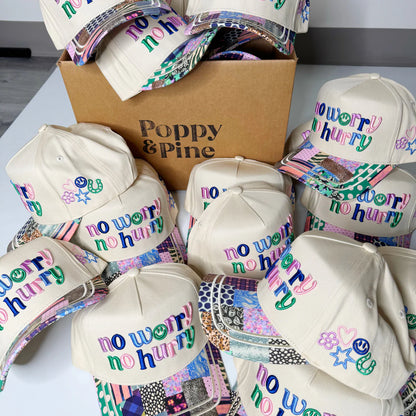 White hats with 'no worry no hurry' text and colorful designs on a white surface with a Poppy & Pine box.