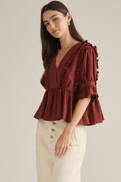 A woman modeling a rust-brown ruffled blouse with elbow-length sleeves and cream-colored pants, photographed against a plain light gray studio background.