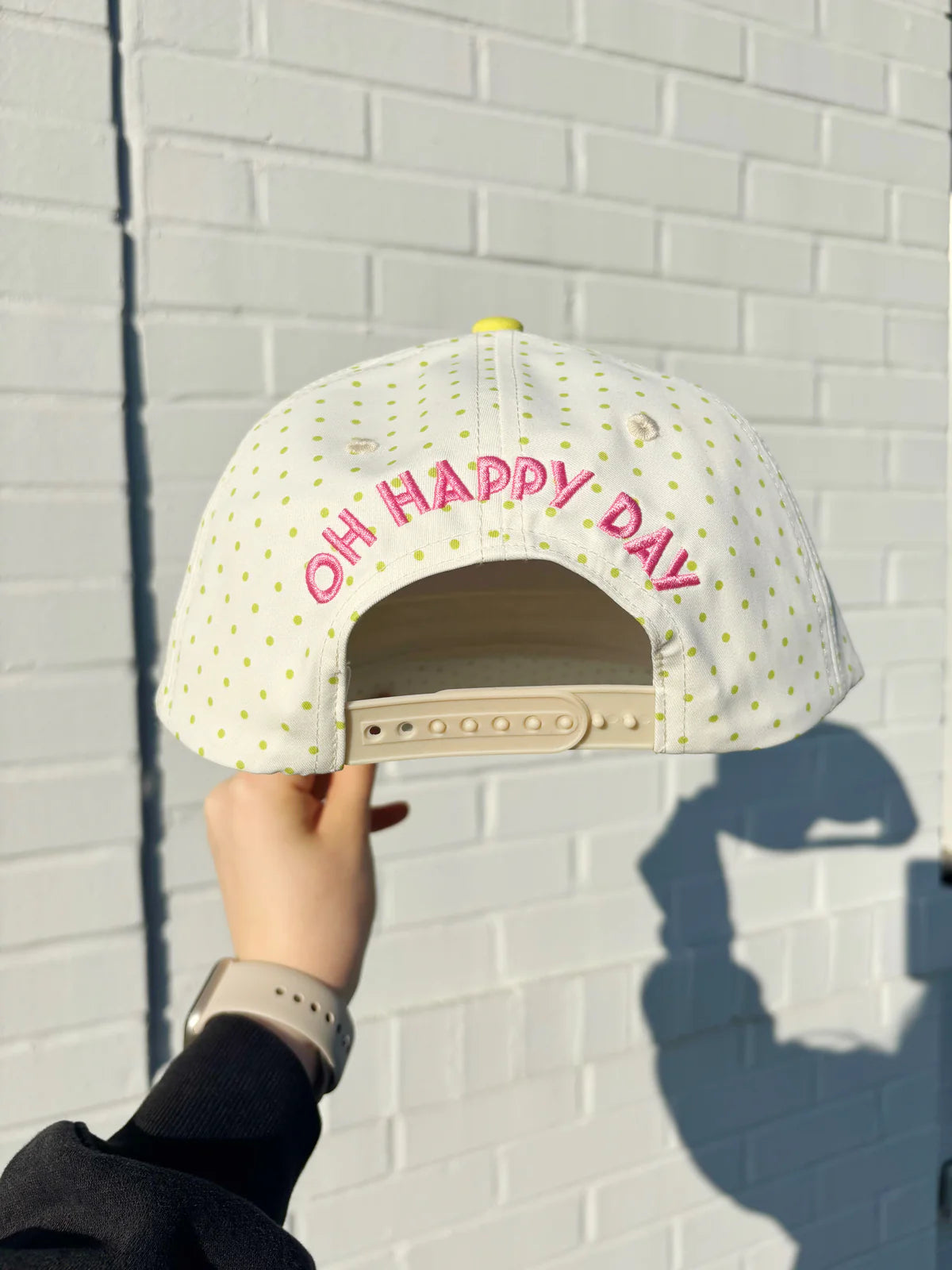 White cap with 'OH HAPPY DAY' text held against a white brick wall