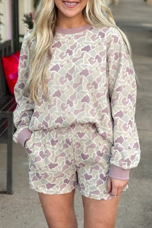 A woman wearing a matching long-sleeve sweatshirt and shorts lounge set with an abstract organic pattern in dusty pink, mauve, and cream, photographed outdoors on a sidewalk.