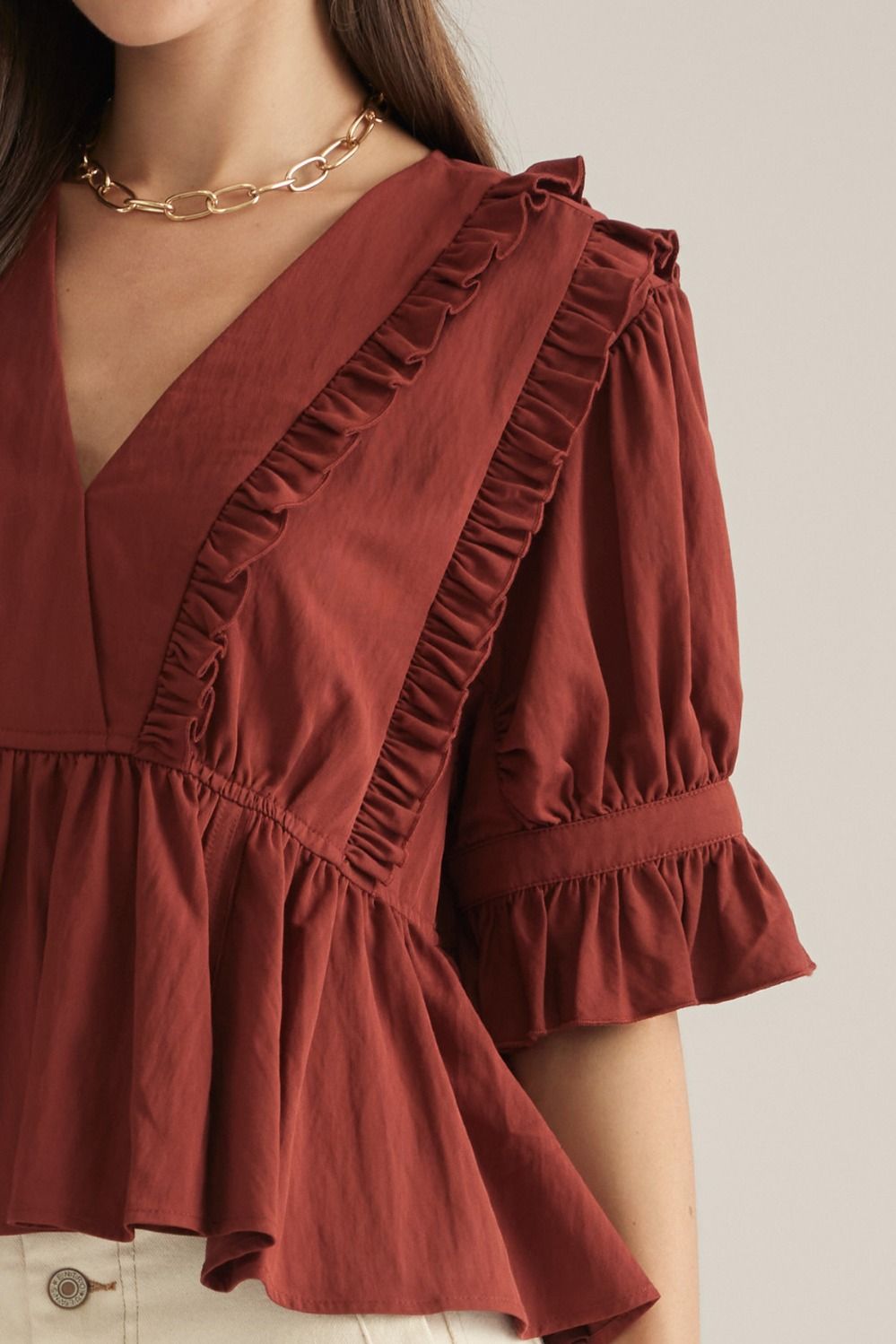 A close-up of a woman wearing a rust-colored blouse with ruffle details, puff sleeves, and a gathered peplum hem, paired with a gold chain necklace.
