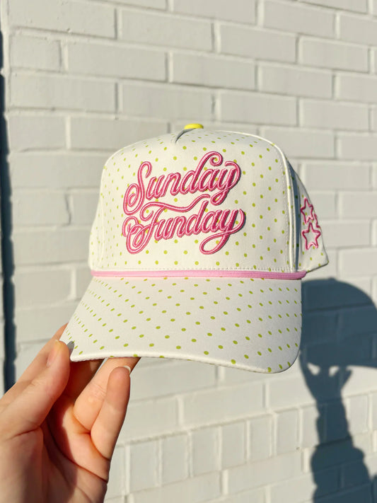 White cap with 'Sunday Funday' text held against a light brick wall.