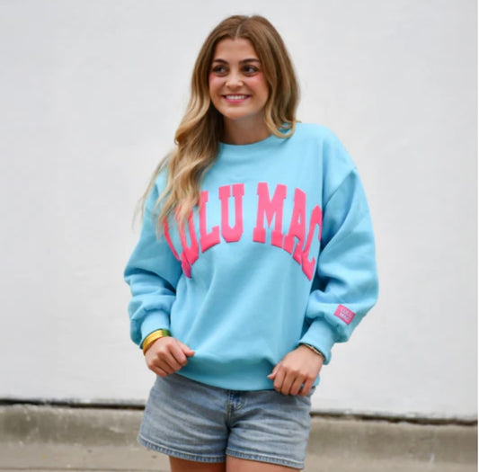 A woman wearing a light blue crewneck sweatshirt with 'LULU MAC' printed in pink across the chest, paired with denim shorts, standing against a plain white wall.