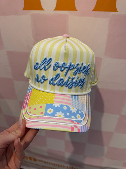 Colorful striped cap with 'all oopsies no daisies' text held against a pink checkered background