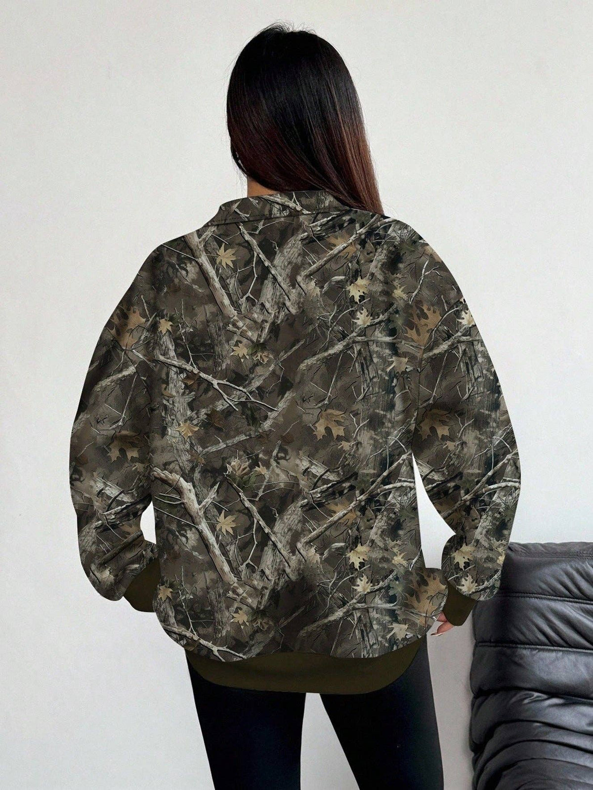 A person shown from the back wearing a camouflage jacket with a woodland tree-bark and leaf pattern in shades of brown, green, and gray, paired with black pants.