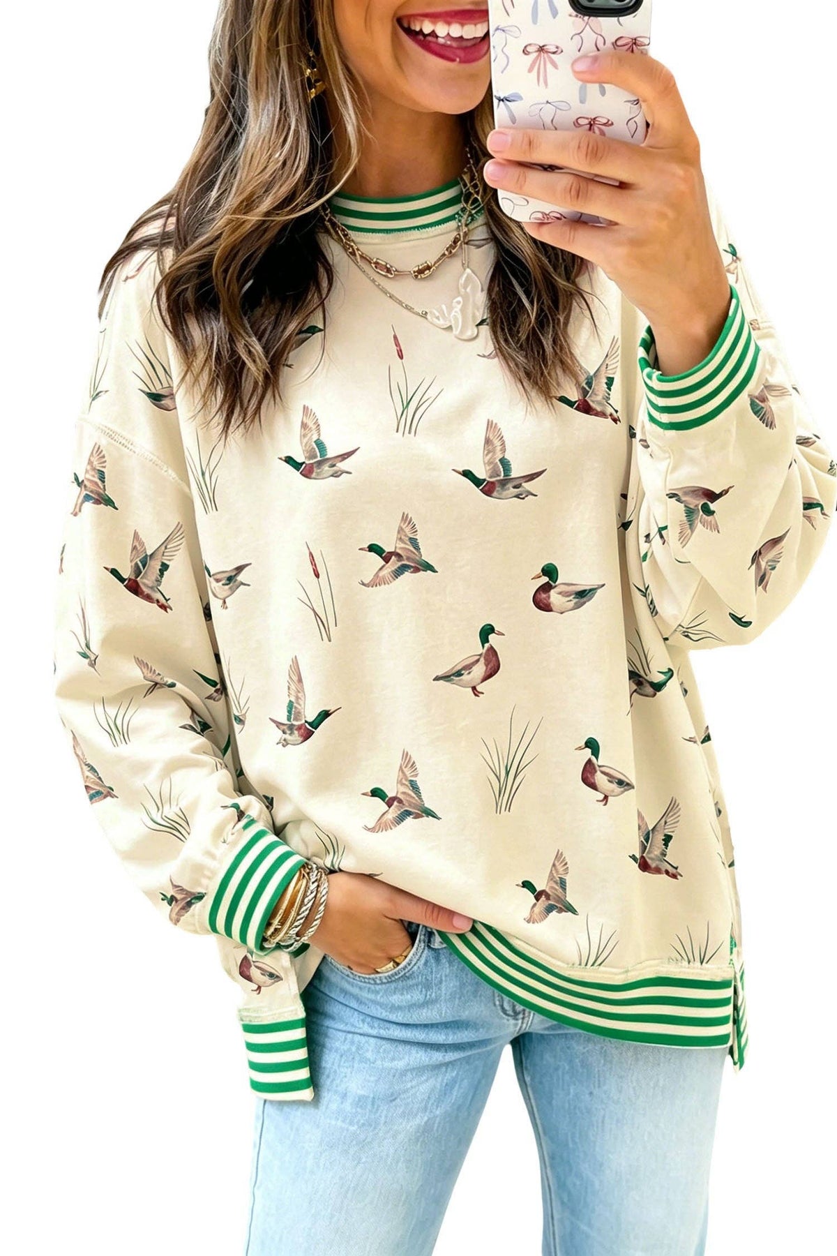 A woman takes a mirror selfie wearing a cream-colored long-sleeve sweatshirt with a green and brown duck hunting pattern and green striped ribbed cuffs and hem, paired with light blue jeans.